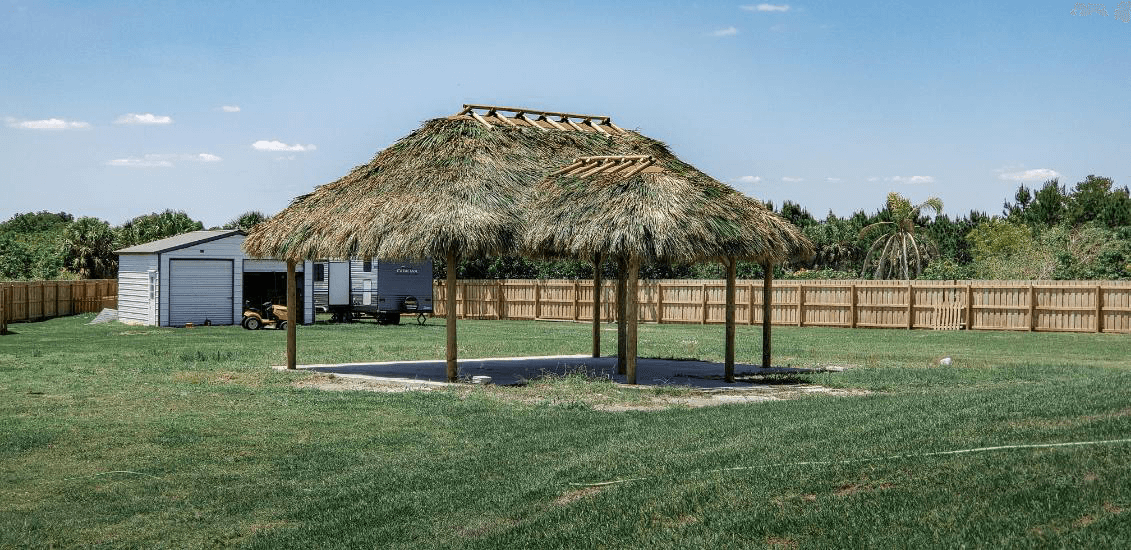 A thatched-roof structure stands in a grassy yard. A wooden fence and a small building are in the background under a blue sky.