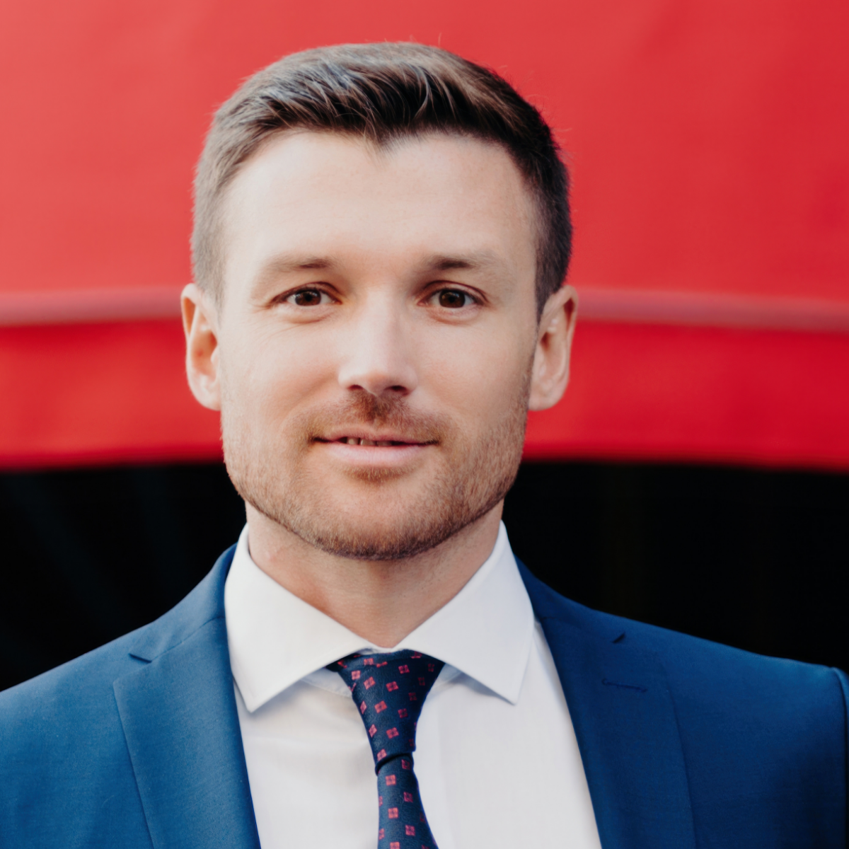 Man in blue suit, white shirt, and patterned tie smiles in front of a red backdrop.