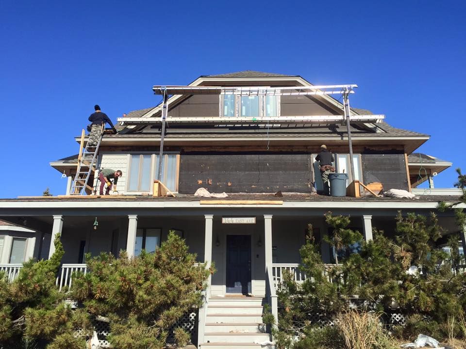 Construction workers on a roof, removing shingles. House is light gray with brown trim. Blue sky.