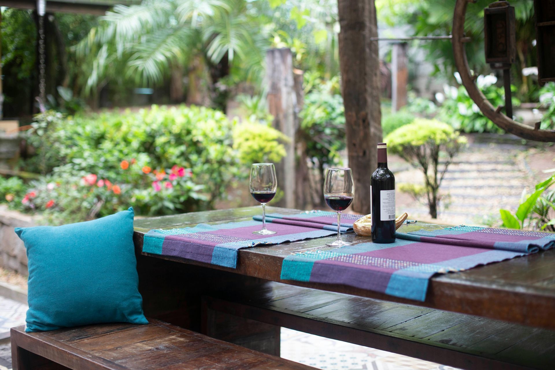 Wooden table set with wine, glasses, and a blue pillow, in a lush outdoor garden setting.
