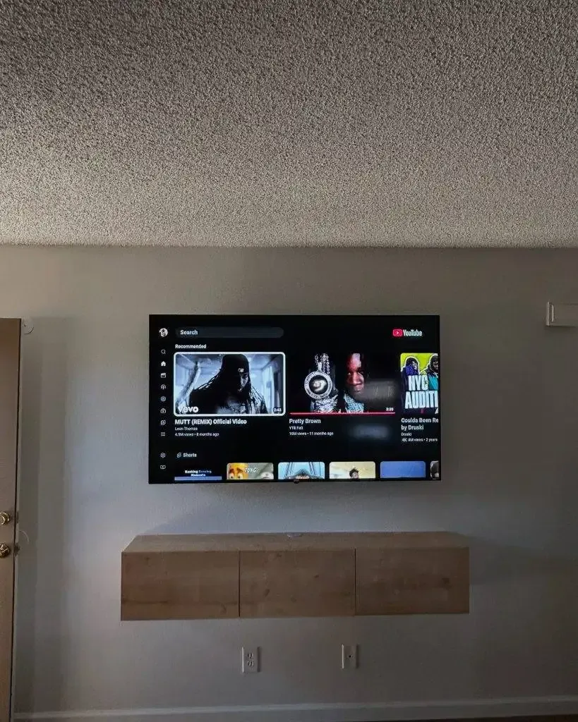 A wall-mounted television displaying a YouTube interface above a light wood floating shelf on a plain gray wall.