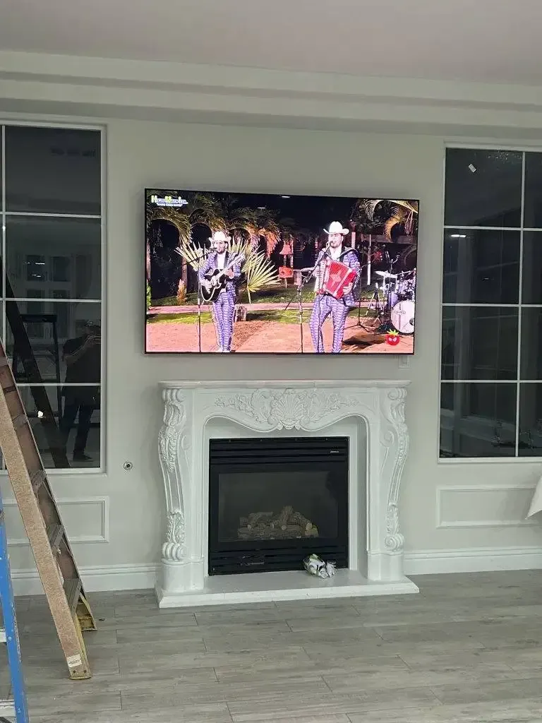 A flat-screen television mounted above an ornate, white carved fireplace in a modern, light-filled living room.