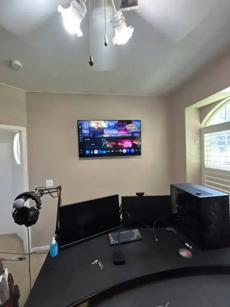 A desk setup with two monitors, a gaming PC, and a mounted TV in a room with a light-colored wall and overhead light.