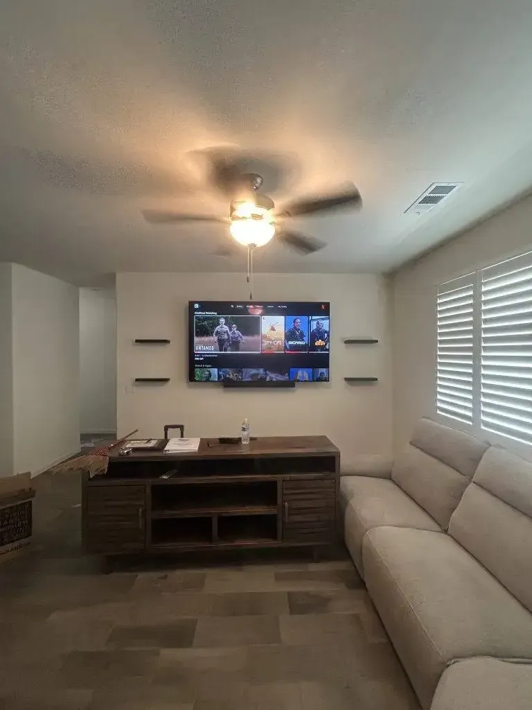 A television mounted above a wooden media console in a living room with a neutral-toned sofa and a ceiling fan.