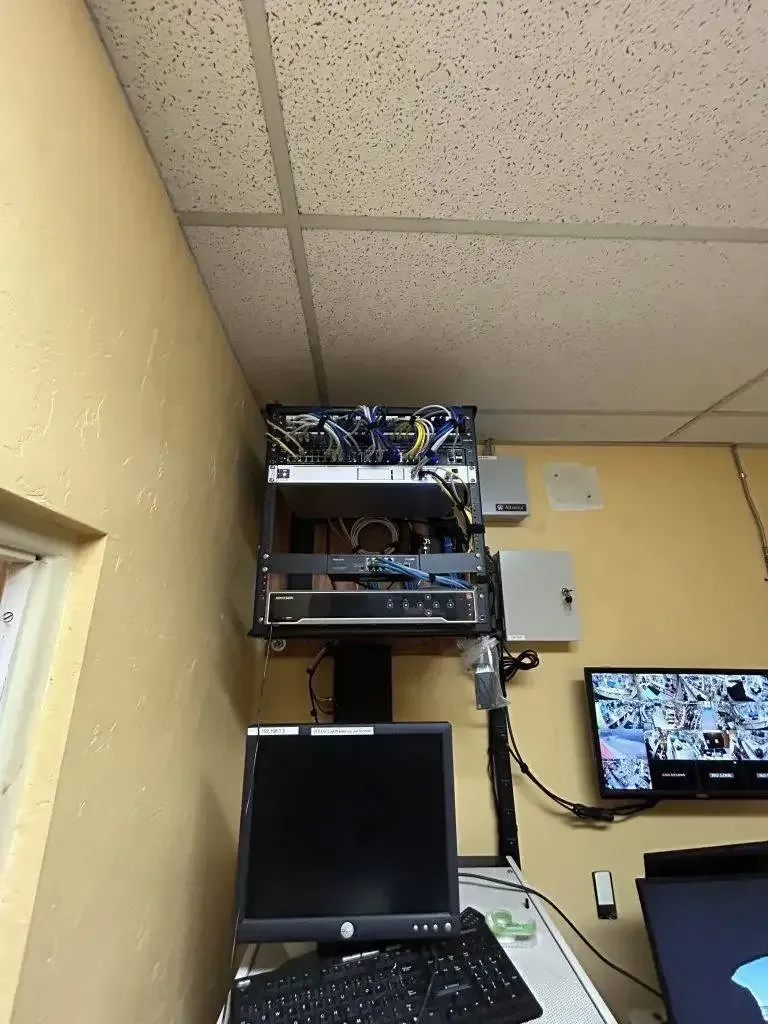 A server rack mounted high on a tan wall above a computer workstation with monitors in a small office setting.