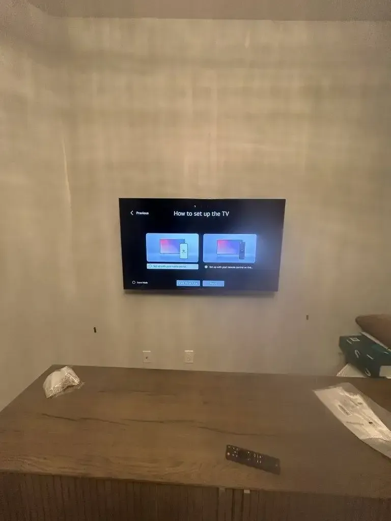 A television mounted on a beige wall displays an Apple TV setup screen above a dark wooden desk.