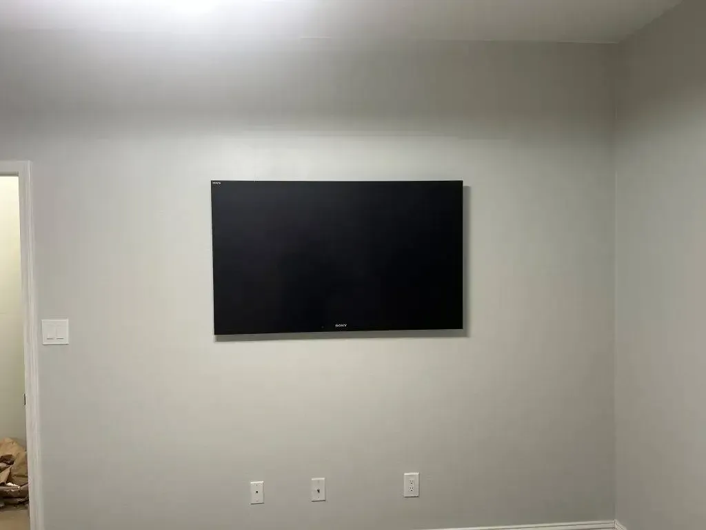 A flat-screen television mounted on a plain, light-gray wall above three electrical outlets.