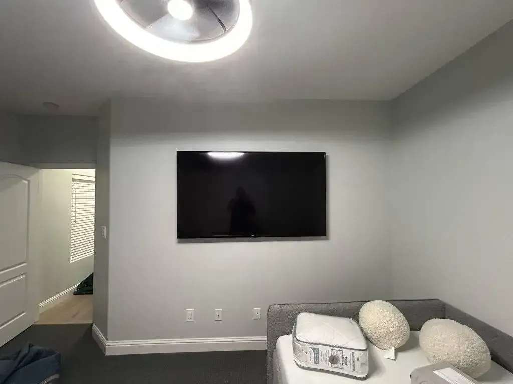 A mounted flat-screen TV on a light gray wall above a bed with white pillows in a neutral-colored bedroom.