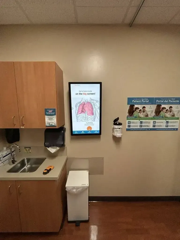 A medical exam room with a sink, a digital display showing a lung diagram, a trash can, and a patient information poster.