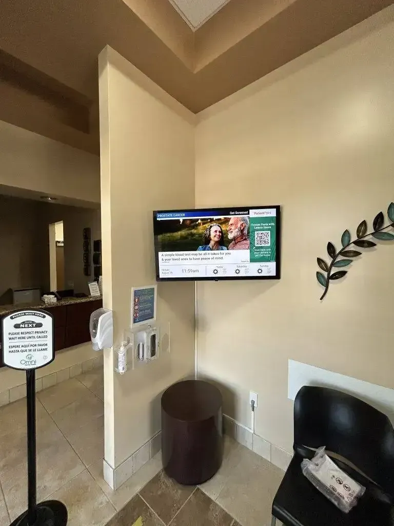 A wall-mounted TV in a waiting area displaying a news broadcast, next to a sanitizer dispenser and a black chair.