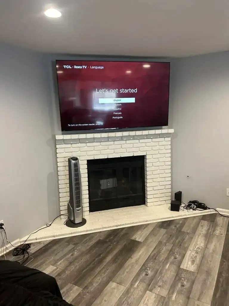 A flat-screen TV mounted above a white brick fireplace in a corner of a room with grey walls and wood-patterned flooring.