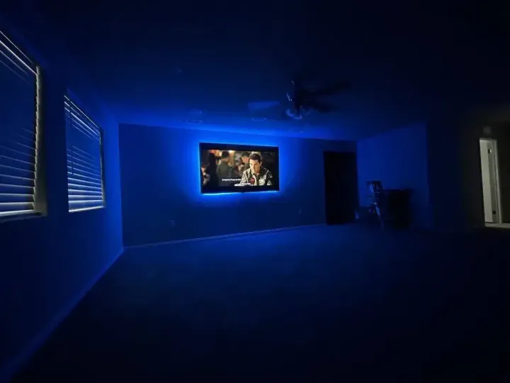 A dark room with a TV mounted on a wall, illuminated by vibrant blue LED bias lighting, with two windows on the left.