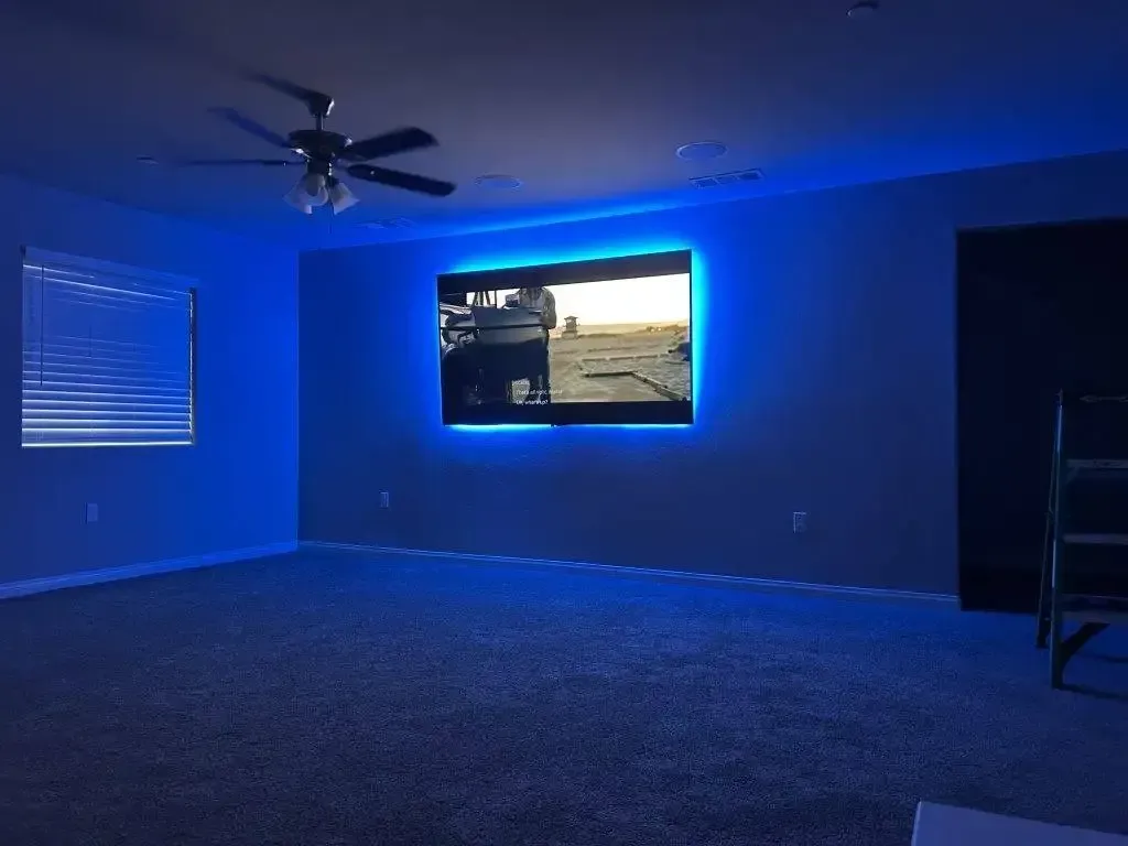 A wall-mounted TV framed by vibrant blue LED backlighting in a dim, carpeted room with a ceiling fan.
