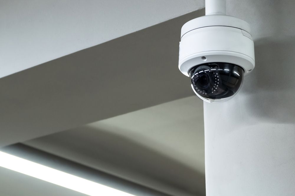 A white security dome camera mounted on a ceiling corner in an indoor setting.