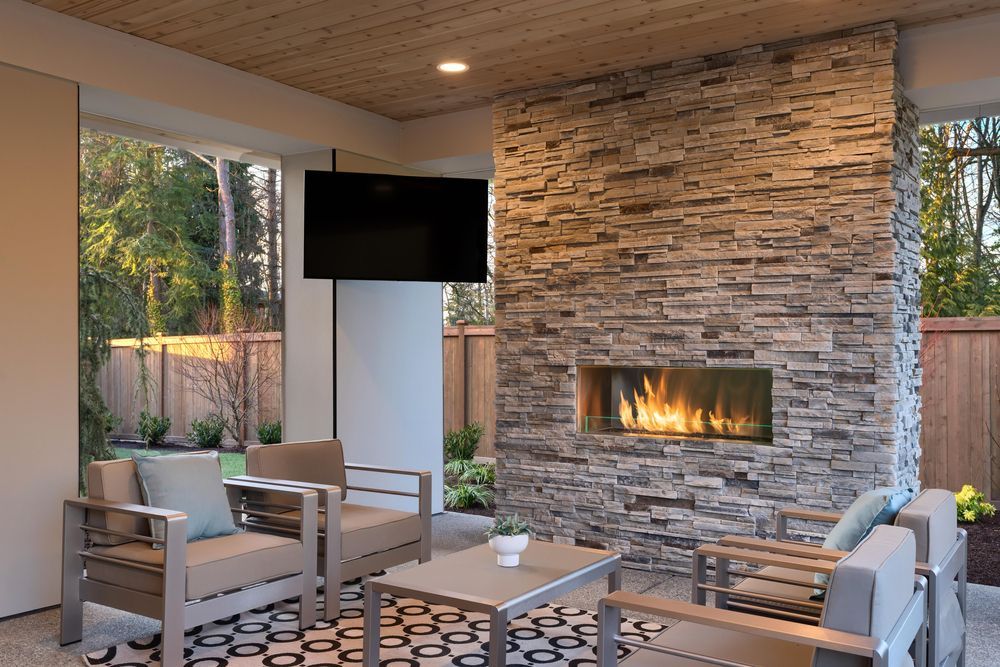 A covered outdoor patio featuring a stacked stone fireplace with a built-in fire, a flat-screen TV, and modern furniture.