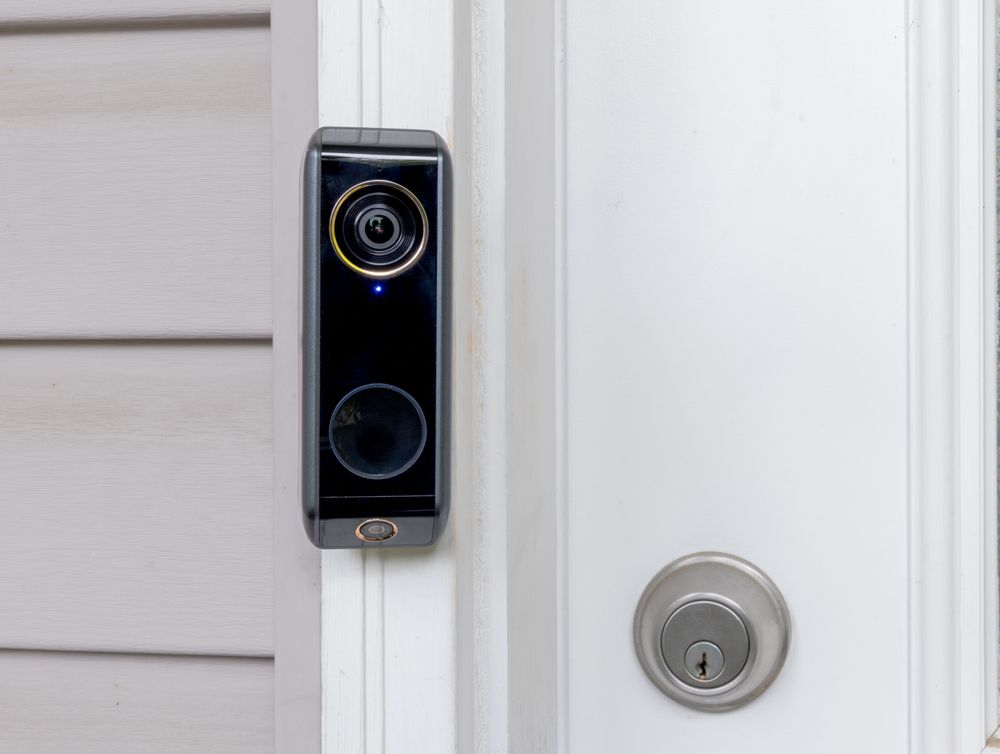 A black smart video doorbell mounted on a white door frame next to a circular silver deadbolt lock.