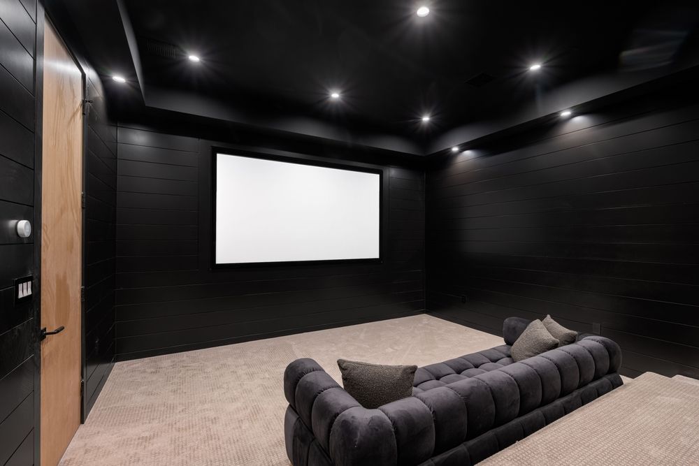 A home theater room with black walls, a large projection screen, recessed lighting, and a long tufted grey sofa.