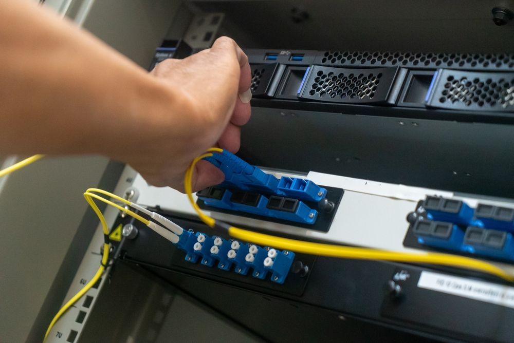 A hand connects a yellow fiber optic cable into a blue port on a server rack.
