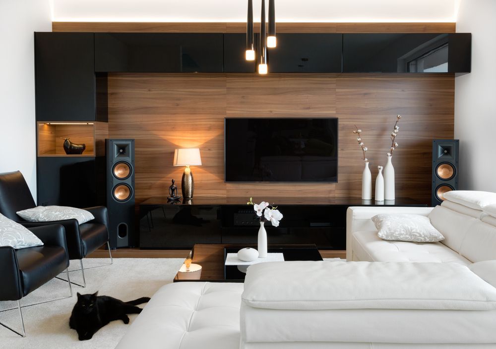 A modern living room with a black and wood media wall, a white sectional sofa, black chairs, and a black cat on the floor.