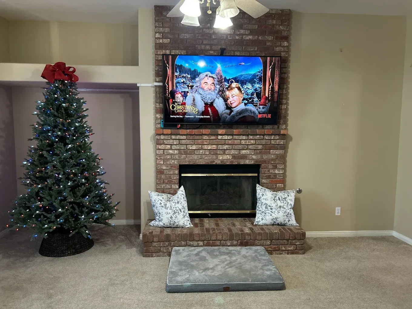 A lit Christmas tree stands in a room next to a brick fireplace with a mounted television and two patterned pillows.
