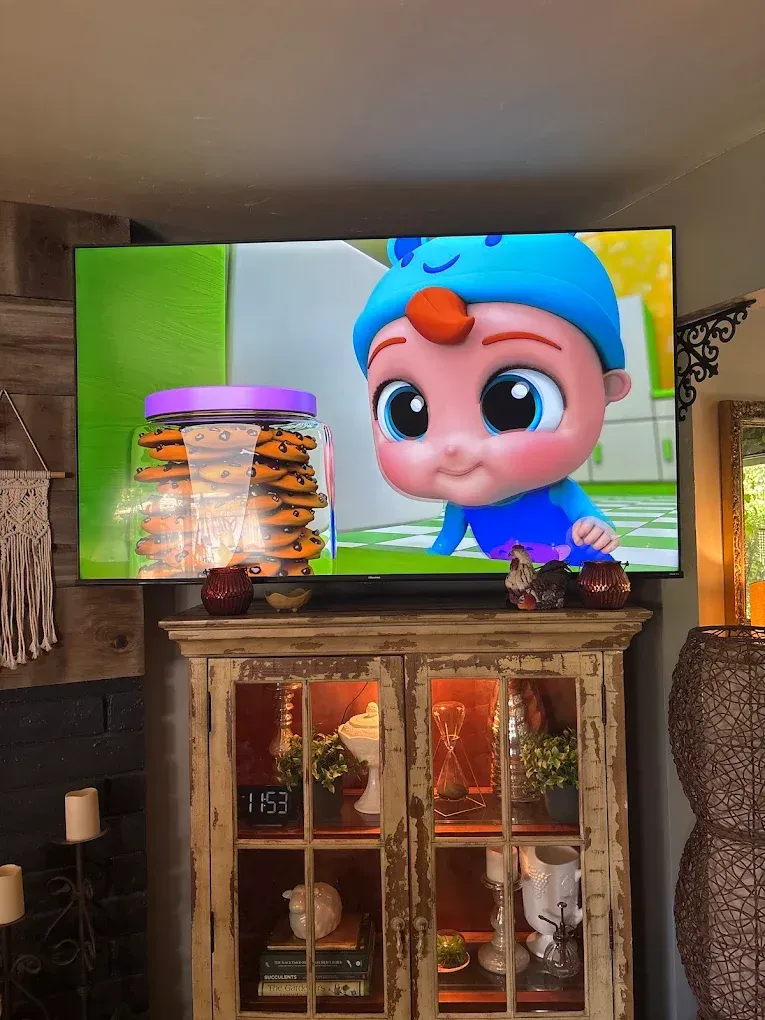 A television screen displays a Cocomelon character near a jar of cookies, positioned above a rustic wooden display cabinet.