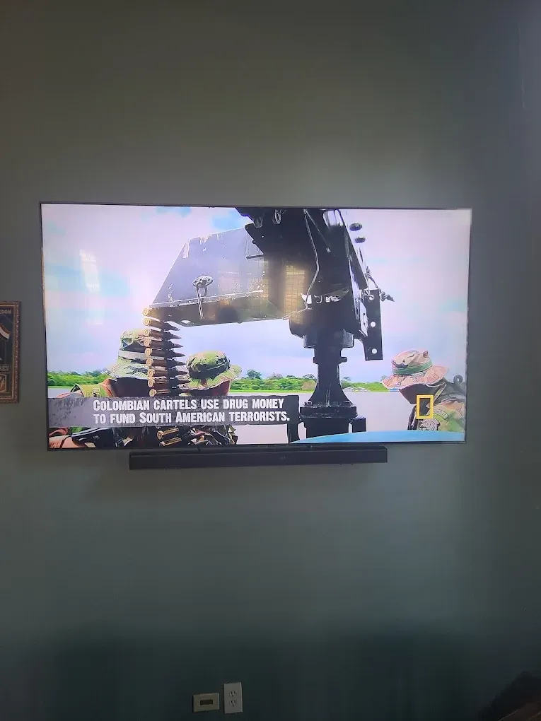 A television screen shows soldiers patrolling a river on a boat with a mounted machine gun, with a National Geographic logo.