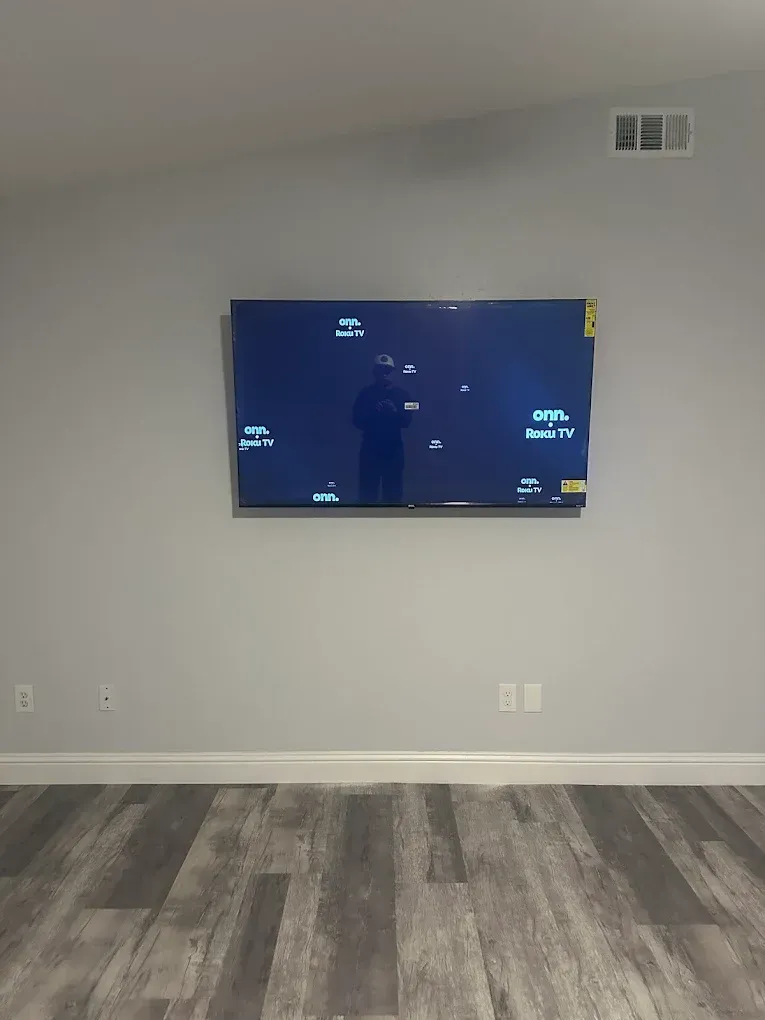 A flat-screen television mounted on a plain light gray wall in an empty room with wood-grain flooring.