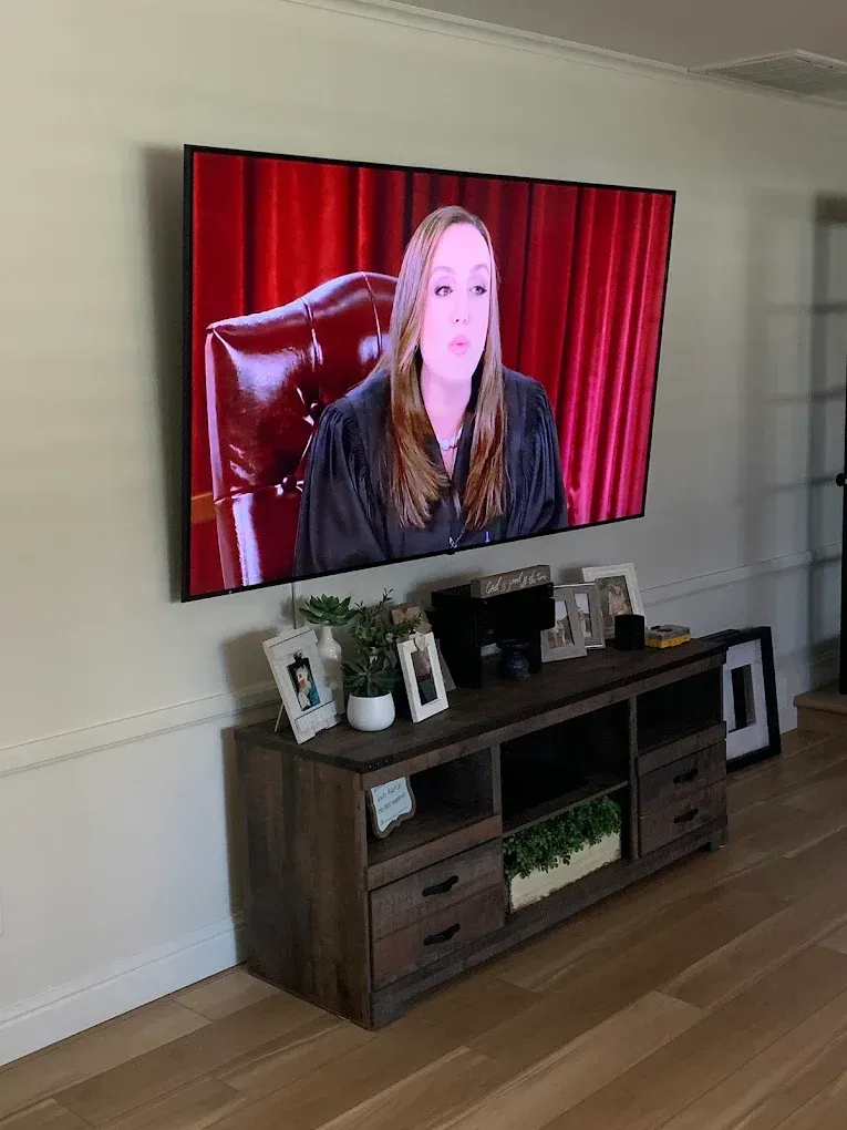 A large flat-screen TV mounted on a wall displays a person in a judge’s robe against a red background above a wooden console.