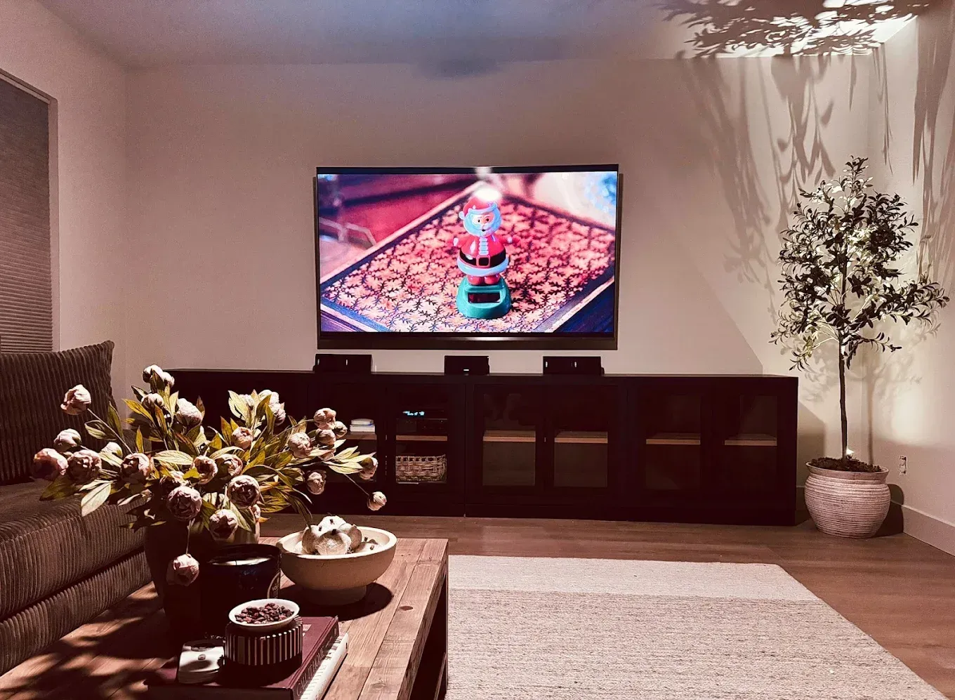 A living room with a large TV mounted above a long dark wooden cabinet, a coffee table with decor, and a potted tree.