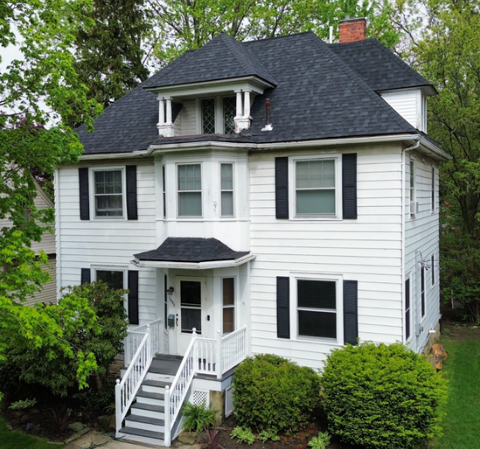 White two-story house with dark roof, black shutters, front porch, and white steps surrounded by green trees and shrubs