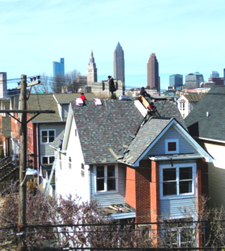 Rooftop houses in foreground with a city skyline and tall towers in the background under a clear sky