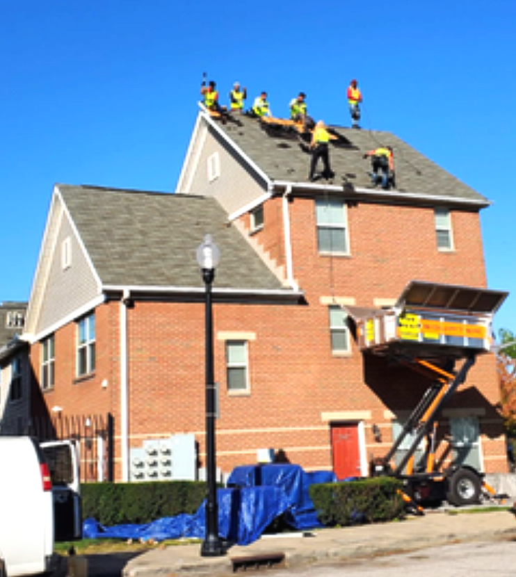 Workers repair a roof on a red brick house while a lift raises materials at the front.