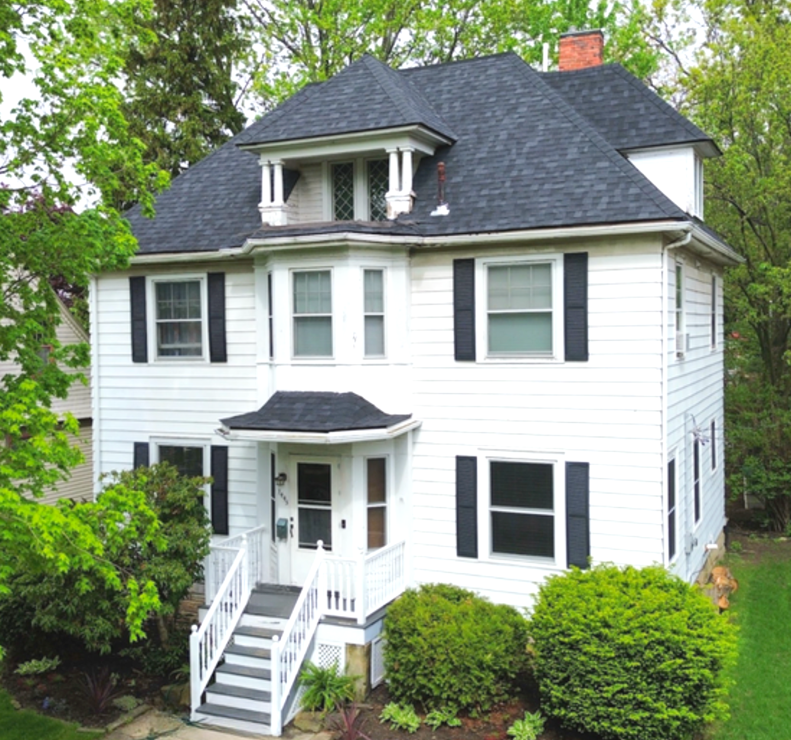 White two-story house with black shutters, front porch, and steps surrounded by green trees and shrubs