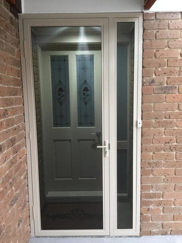 Beige screen door and side panel in front of a white door with decorative glass, set in a brick entryway.