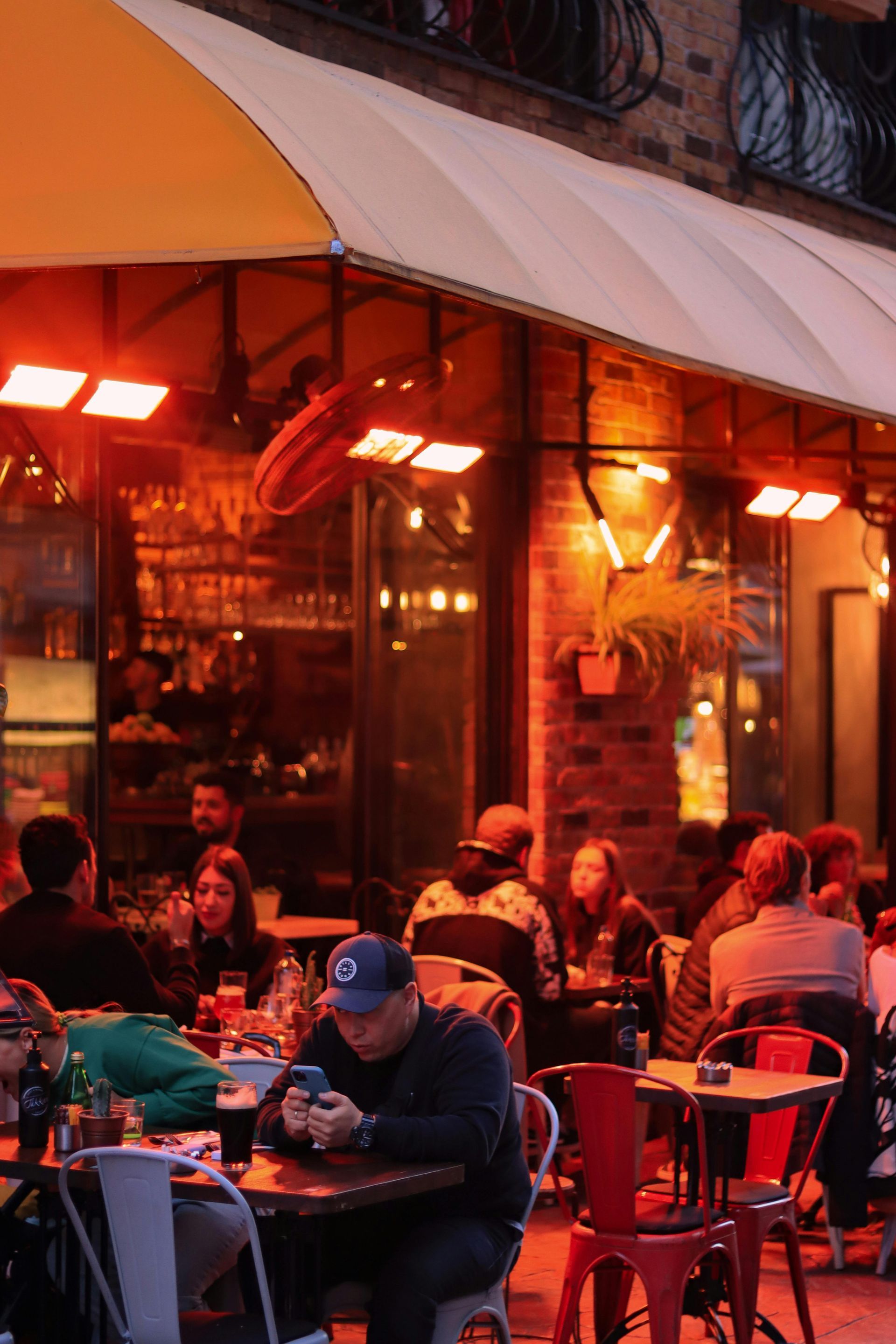 Outdoor café with people seated at tables under warm red lights and an awning at dusk
