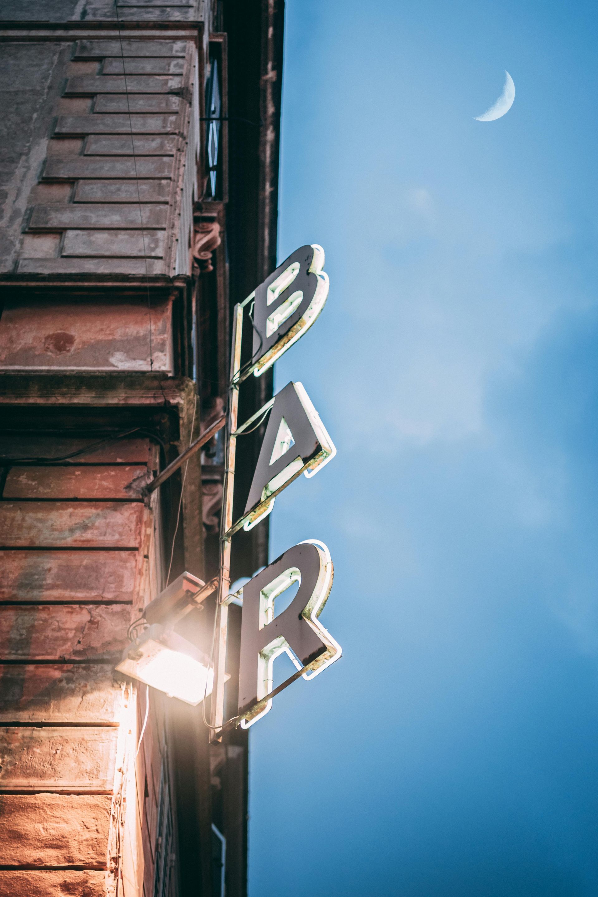 Vertical “BAR” sign on a wooden building at dusk, with a crescent moon in a blue sky.