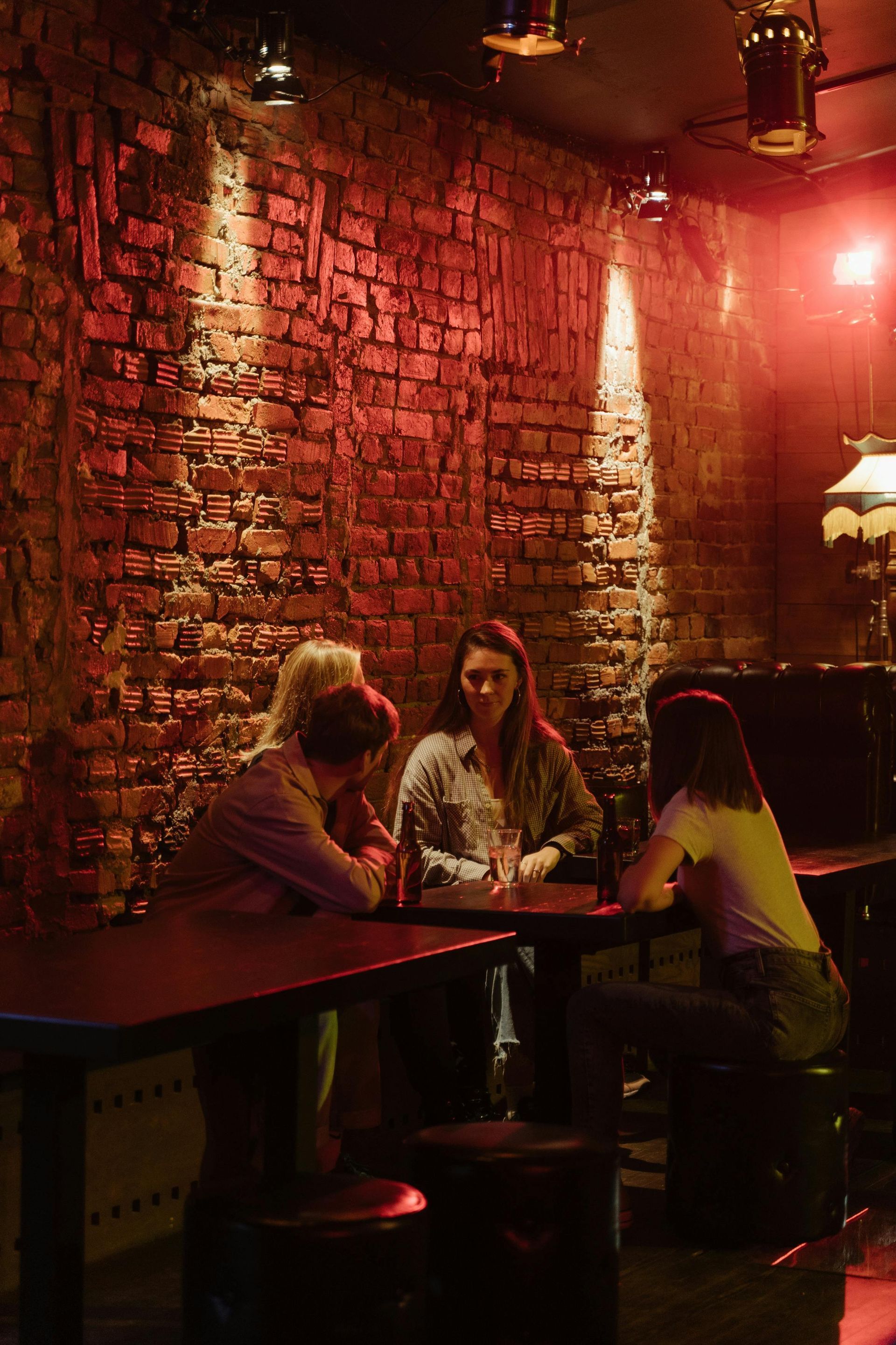 Three people sit at a table in a dim, red-lit brick-walled bar