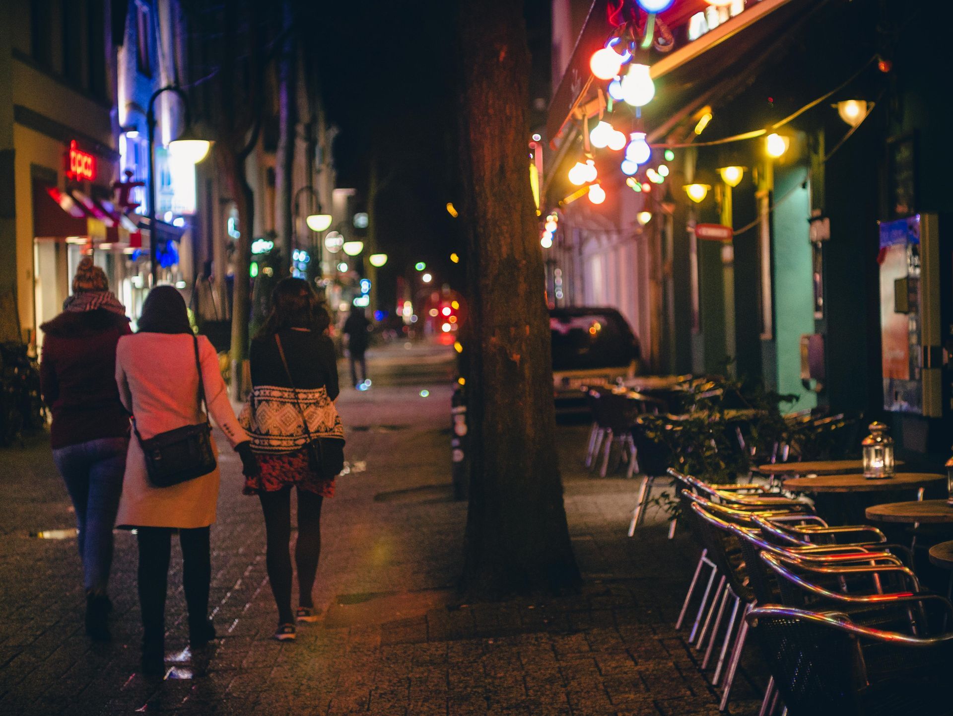People walking down a neon-lit street at night past café tables and storefronts