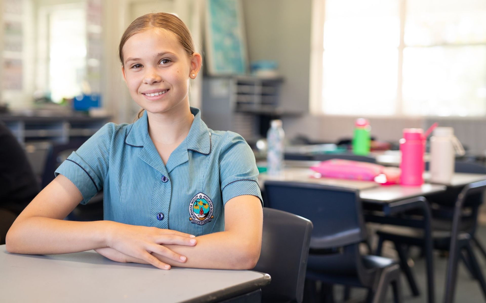 Girl in school uniform smiles at desk in classroom.