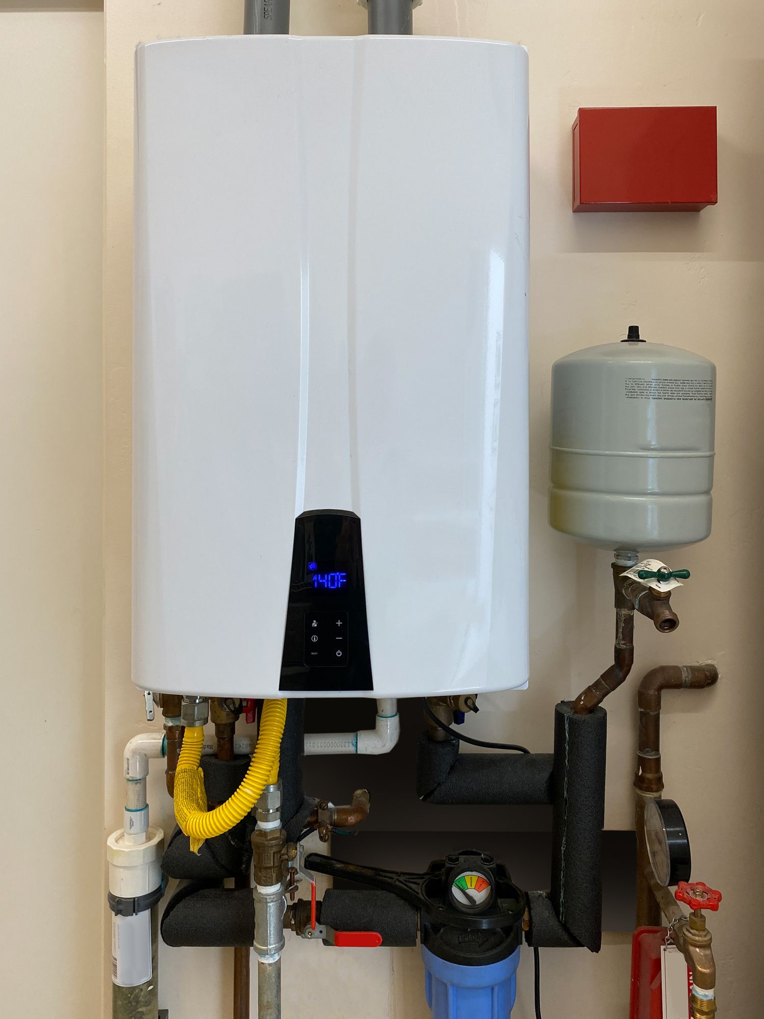 Wall-mounted white tankless water heater with pipes and other components, mounted on a beige wall.