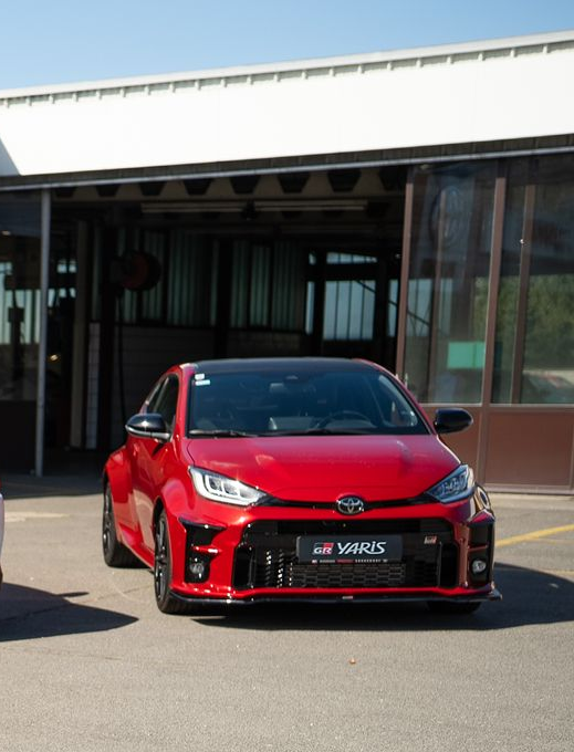 Roter Toyota GR Yaris mit schwarzem Dach, an einem sonnigen Tag in der Nähe einer Garage geparkt.