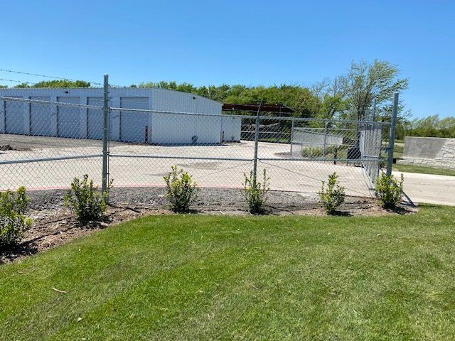 A chain link fence surrounds a parking lot with a white building in the background.
