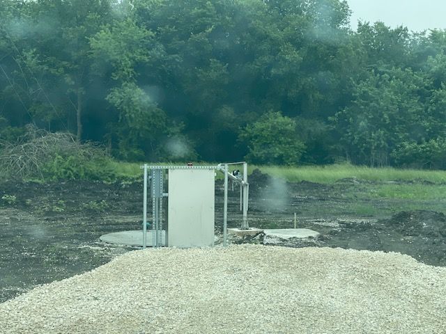 A large pile of gravel is in the middle of a field with trees in the background.