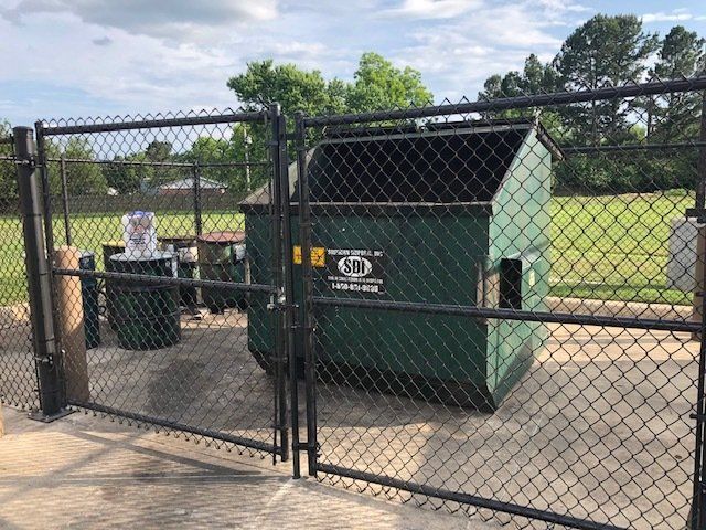 A green dumpster is behind a chain link fence.