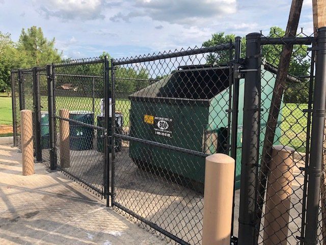 A green dumpster is behind a chain link fence.
