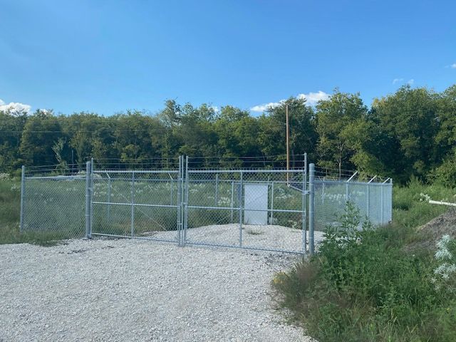 A metal fence is surrounding a gravel area with trees in the background.