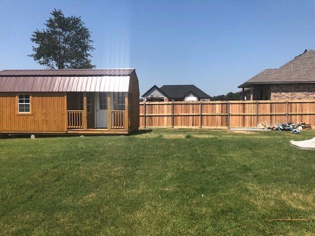 A wooden shed is sitting in the middle of a grassy yard next to a wooden fence.