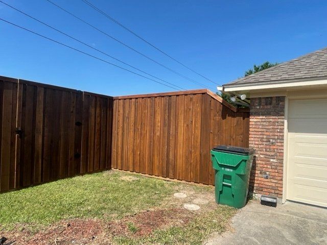 A green trash can is sitting in front of a wooden fence.