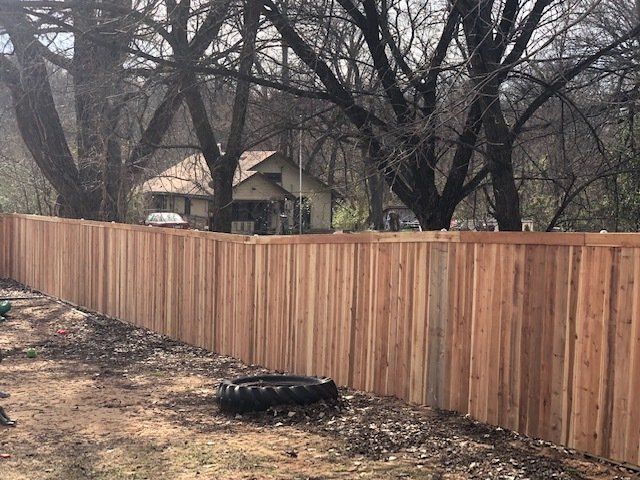 A wooden fence surrounds a yard with trees and a house in the background.