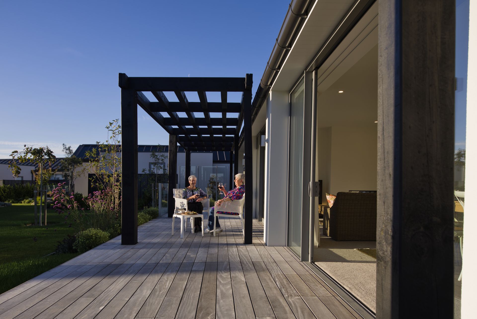 Modern patio with wooden deck, black pergola, and people dining beside large open glass doors at sunset